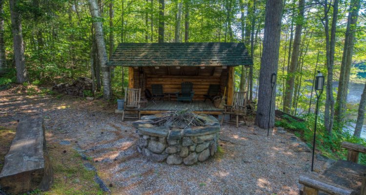 A log cabin-style lean-to sits behind a stone fire pit waiting for visitors to cozy up next to it